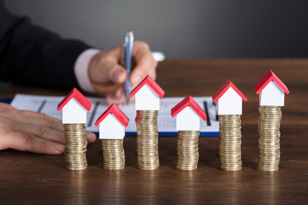 A hand signing a document with small house models placed on stacks of coins in the foreground, symbolizing real estate taxes in Beaufort County, South Carolina.