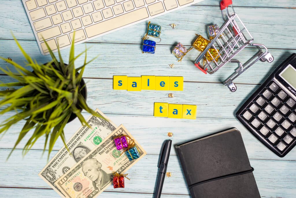 Flat lay of sales tax concept with a calculator, miniature shopping cart, dollar bills, and letter blocks spelling 'sales tax' on a wooden background.